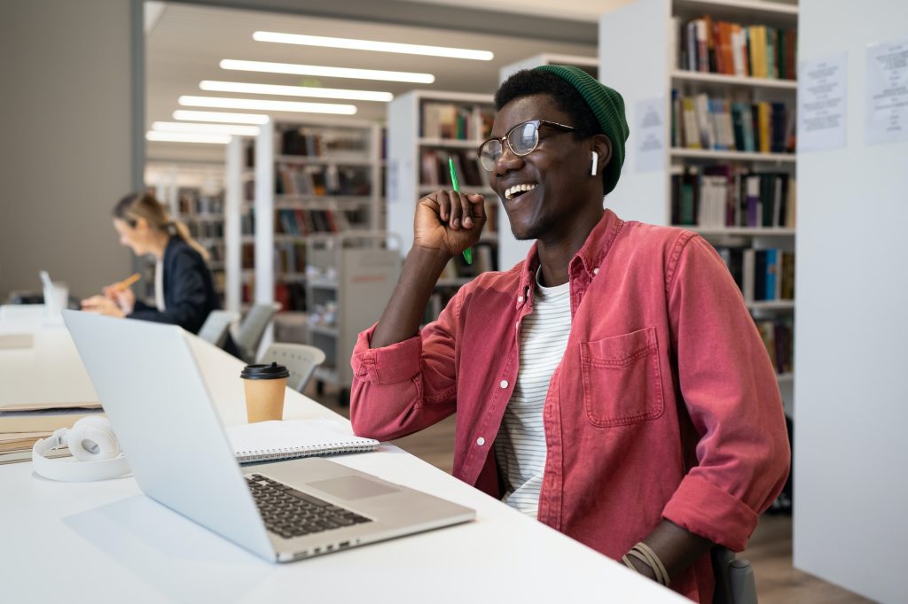 Happy excited African American man student sits in library at table with laptop and laughing